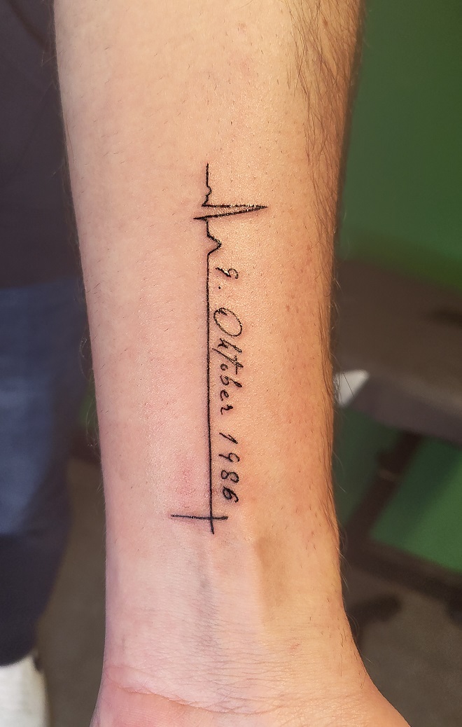 Tattoo eines Schriftzugs mit Datum auf einem EKG-Herzschlag auf dem Unterarm