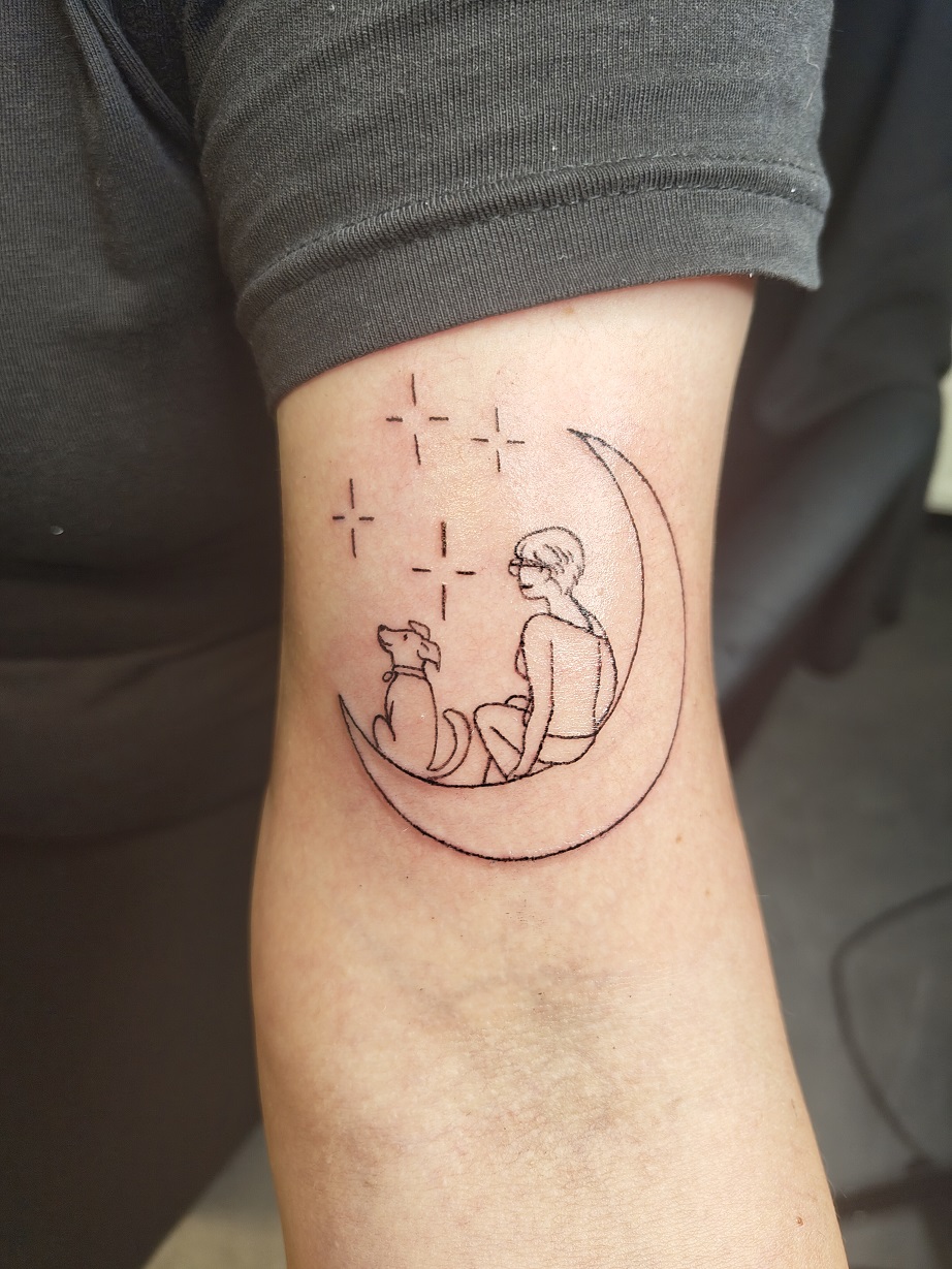 Tattoo eines Mondes auf dem eine Frau und ein Hund sitzen und in die Sterne schauen auf dem inneren Oberarm
